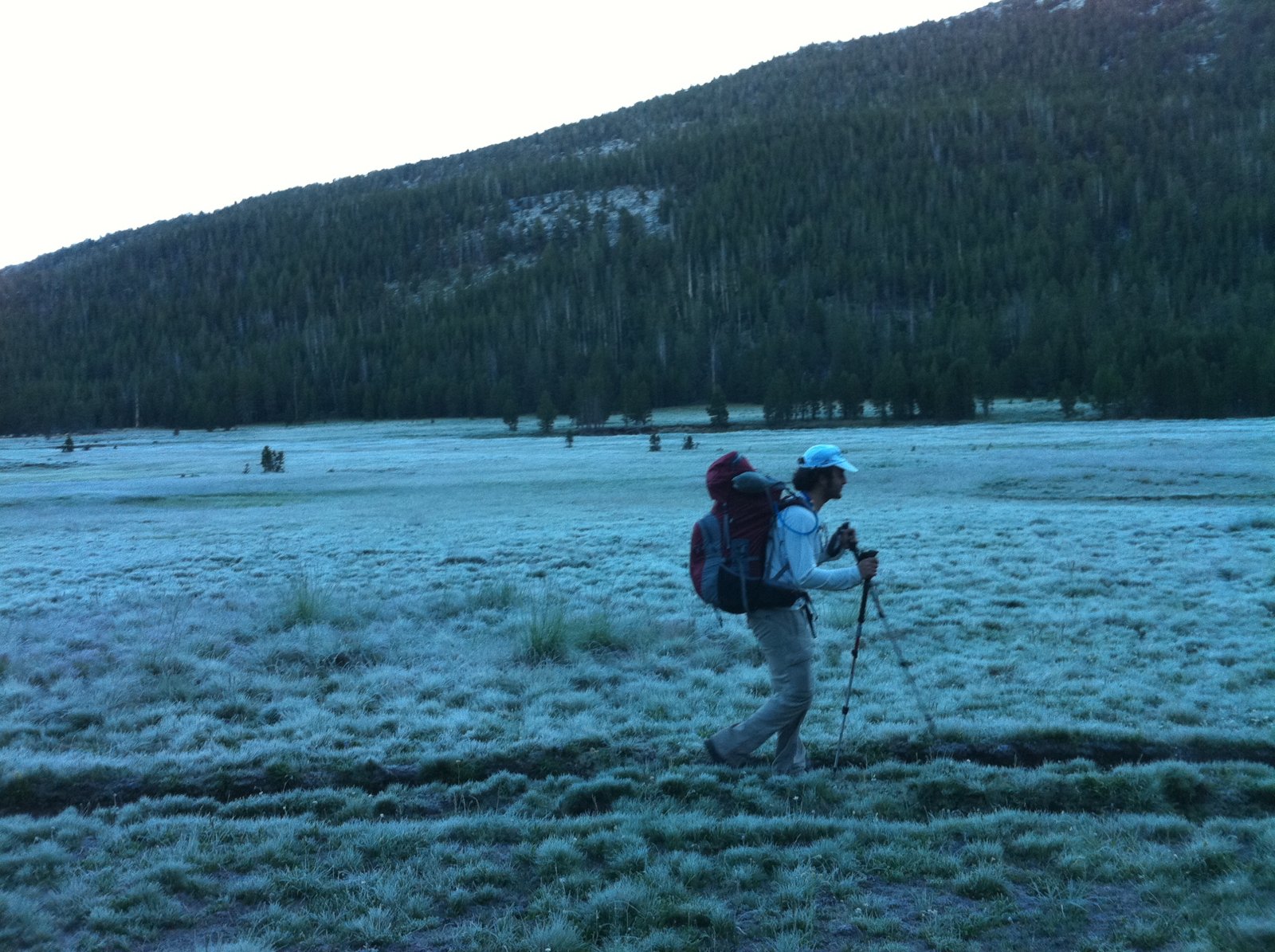 A backpacker leans into a climb with two trekking poles across a frosty Sierra meadow at dawn