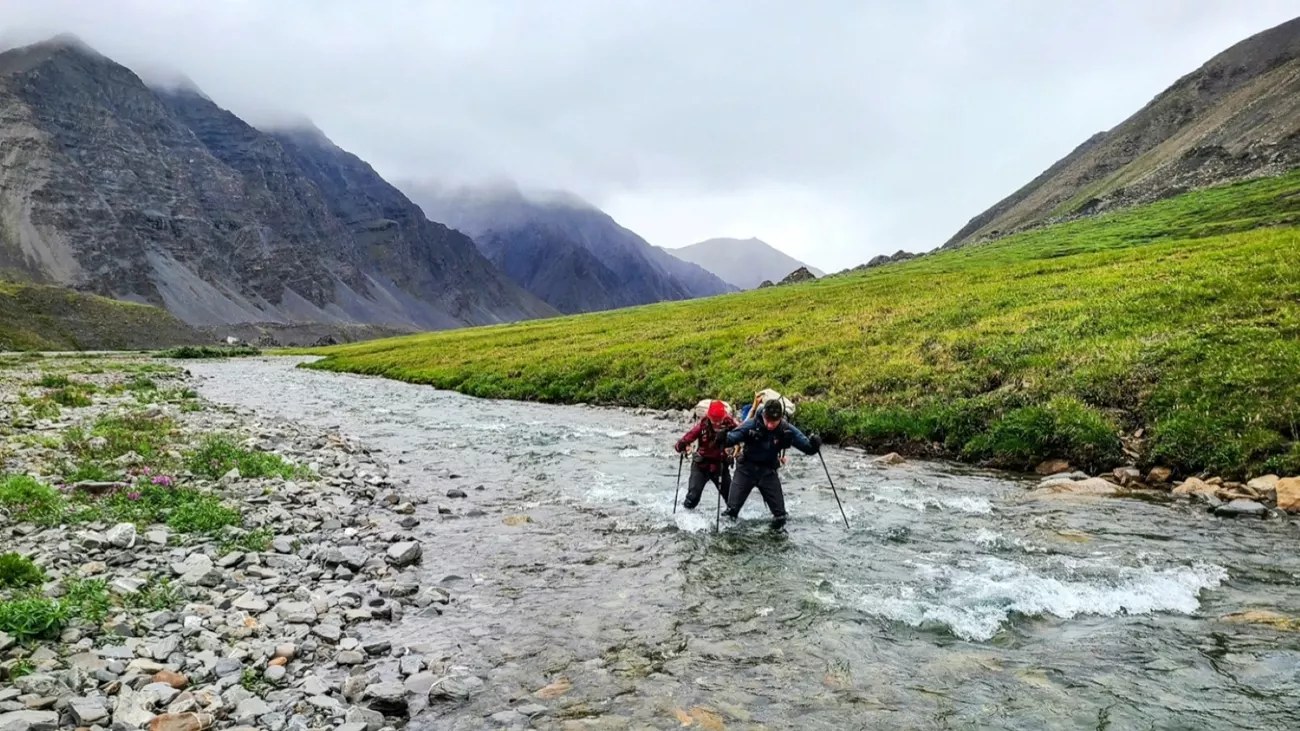 Two backpackers with trekking poles face upstream as they ford a cold, fast river below cloud-capped mountains